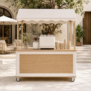 Könnte beinhalten: Ein mobiler Kaffeewagen mit weißem, gewelltem Vordach und hellen Holzpaneelen. Auf der Theke stehen eine Glasvase mit weißen Blumen, eine weiße Kaffeemühle, eine weiße Espressomaschine und mehrere Stapel beigefarbener Pappbecher für Kaffee.