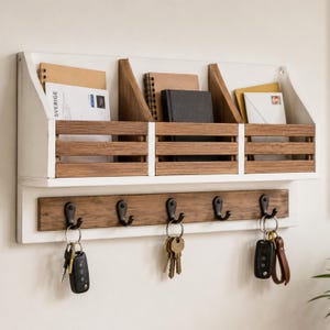 Peut inclure: Un organisateur mural composé de trois compartiments de rangement en bois avec des cadres blancs et des lattes en bois naturel. Sous les compartiments se trouve une planche horizontale en bois avec cinq crochets en métal noir portant trois trousseaux de clés.