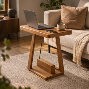 Puede incluir: Una mesa auxiliar en forma de C hecha de madera de color marrón claro, con un tablero rectangular, una pata de soporte vertical y una base con un bloque de almacenamiento horizontal integrado. Una varilla metálica negra une el tablero a la base.