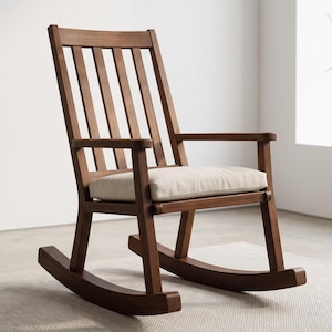 May include: A wooden rocking chair with a medium brown finish, featuring a high backrest with five vertical slats, two armrests, and a thick, off-white upholstered seat cushion. The chair is supported by two curved rockers at the base.