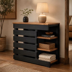 May include: A black wooden console table with a natural wood top and horizontal slat design. The unit features three open shelves on the right side, currently holding a small wooden bowl, a book titled Kinfolk, a woven basket, and a folded throw.