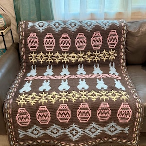May include: A brown crocheted blanket with a repeating pattern of pink Easter eggs, blue bunnies, and yellow and blue geometric shapes. The blanket is draped over a brown leather couch.