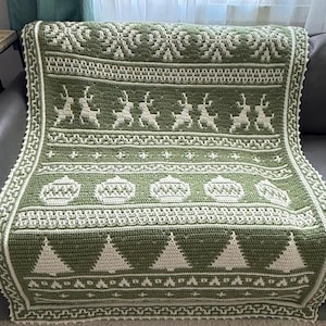 Puede incluir: Una manta rectangular de ganchillo con un diseño bicolor en verde oliva y crema. El patrón presenta bandas horizontales con motivos repetidos de copos de nieve, renos, adornos decorativos y árboles de pino con un borde texturizado.
