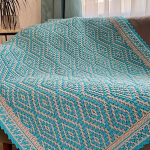 Pode incluir: Um cobertor de crochê feito à mão em tons de azul-turquesa, bege e cinza claro, sobre um sofá. O cobertor apresenta um padrão geométrico de diamantes e uma borda ondulada. Um item de decoração aconchegante.