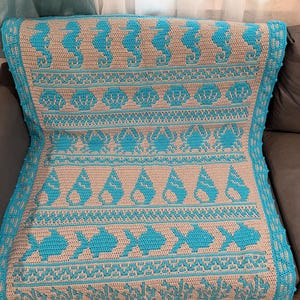 May include: A crocheted blanket with a marine theme. The blanket has a light beige background with turquoise sea creatures, including seahorses, seashells, crabs, and fish. The border is turquoise. The blanket is folded over a sofa.