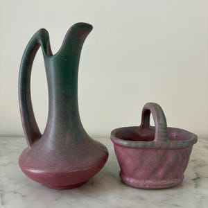 May include: A two-piece ceramic set featuring a tall, slender pitcher with a curved handle and a small basket with a handle. The pitcher is a gradient of green to pink, while the basket is pink. Both pieces have a textured surface.