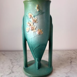 May include: A teal ceramic vase with a flared top and a round base. The vase features a floral design with light yellow and pink accents. The vase has two vertical supports on either side.
