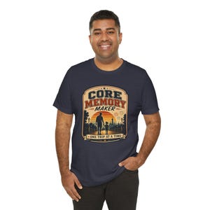 May include: A navy blue short-sleeve t-shirt featuring a graphic print with the text "CORE MEMORY MAKER" and "ONE TRIP AT A TIME". The design depicts a silhouette of an adult and a child walking toward a sunset with fireworks and park structures.