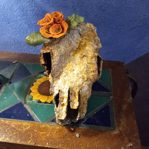 May include: A brown, weathered, animal skull with a cluster of orange paper flowers on top. The skull is sitting on a table with a colorful tiled surface.