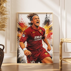 Può includere: Stampa artistica incorniciata di un calciatore in uniforme rossa, con la scritta "INA Assitalia". L'opera presenta una posa dinamica su uno sfondo astratto e colorato. La stampa è esposta in una cornice di legno chiaro.