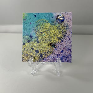 May include: A square card featuring a vibrant abstract watercolor design with splattered yellow, blue, and purple paint. The card is adorned with a cluster of small white pearls and blue crystals in the top right corner and a single dark bead.