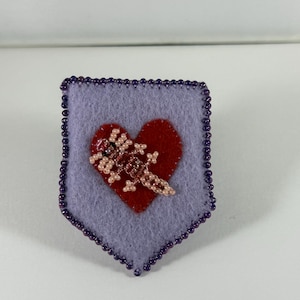 Puede incluir: Un broche de fieltro lavanda hecho a mano con un corazón rojo y un diseño de lagarto con cuentas. El broche está bordeado con pequeñas cuentas moradas. El lagarto está hecho de cuentas rosas y marrones.