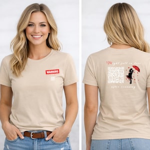 Könnte beinhalten: Beiges T-Shirt mit einem roten "WARNING" Etikett auf der Vorderseite. Die Rückseite des Shirts zeigt einen QR-Code, eine Silhouette eines Paares unter einem roten Regenschirm und den Text "Might fall in love after scanning."