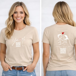Könnte beinhalten: Beiges T-Shirt mit den Worten "You & Me" in Schreibschrift und einem roten Herz. Auf der Rückseite des Shirts befindet sich eine Zeichnung von zwei Strichmännchen, die sich an den Händen halten, mit einem roten Herz darüber. Der Text "one step away" und "from your dream girl" ist ebenfalls vorhanden.