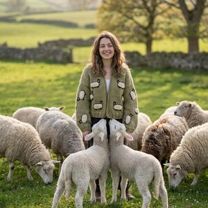 Könnte beinhalten: Eine Frau in einem grünen Cardigan mit Schafapplikationen steht auf einer Wiese mit einer Schafherde. Der Cardigan ist hellgrün mit weißen und schwarzen Schafmotiven. Die Schafe grasen auf grünem Gras.
