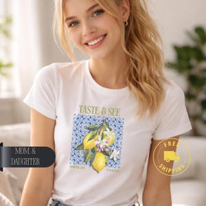 Może przedstawiać: Biała koszulka z ilustracją cytryny i napisem "TASTE & SEE". Projekt zawiera cytryny, kwiaty i liście na niebiesko-białym wzorzystym tle. Koszulka ma również tekst "FRUITFUL LIFE" i "PSALM 34:8".