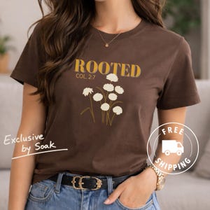 May include: A brown t-shirt with the word "ROOTED" in yellow block letters and the reference "COL. 2:7". Below the text is a graphic of white flowers. The shirt is part of the "Exclusive by Soak" collection.