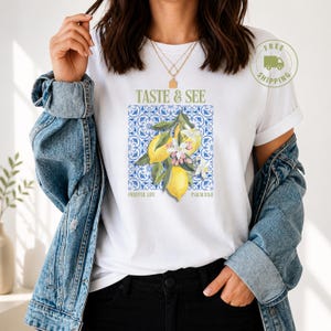 Könnte beinhalten: Ein weißes T-Shirt mit einer Grafik aus gelben Zitronen und weißen Blüten über einem blau-weißen Fliesenmuster. Der Text TASTE & SEE steht über der Grafik, während FRUITFUL LIFE und PSALM 34:8 unter der Illustration aufgedruckt sind.