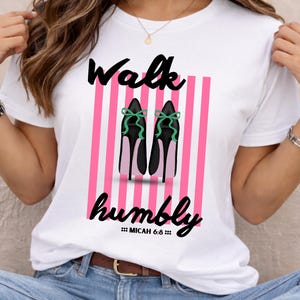 May include: White t-shirt with the words "Walk humbly" in black script, above an illustration of black high heels with green bows. The background features pink and white vertical stripes. The text "Micah 6:8" is below.