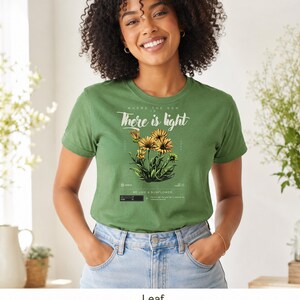 May include: An olive green short-sleeve t-shirt featuring a graphic of yellow sunflowers. The text reads "Where the son is there is light" and "Be like a sunflower" along with smaller decorative elements and a black rectangular graphic detail.