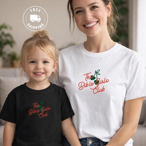 Op de afbeelding: Twee mensen dragen bijpassende t-shirts met de tekst "The Bible Girls Club" in rood script en een groen en rode afbeelding. Eén shirt is zwart, de andere is wit. De afbeelding bevat een "Gratis verzending" logo.