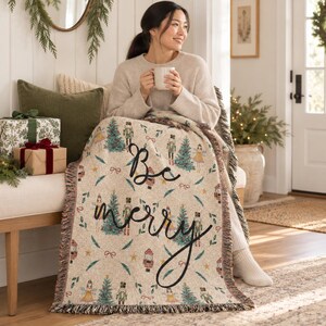 May include: A woven throw blanket featuring a cream-colored background with a repeating pattern of nutcrackers, Christmas trees, and small bows. The text "Be merry" is written in large black cursive script across the center of the blanket.