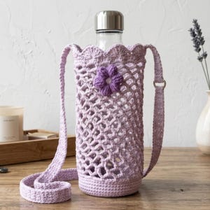 May include: A clear glass water bottle encased in a light purple crocheted holder with a scalloped top edge, a solid base, and a long shoulder strap. A small, dark purple crocheted flower is attached to the front of the open-weave mesh body.