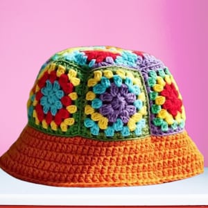 May include: A crochet bucket hat featuring a crown constructed from multi-colored granny squares in shades of red, yellow, blue, purple, and green. The wide, solid brim is worked in a vibrant orange yarn with a dense, uniform stitch pattern.