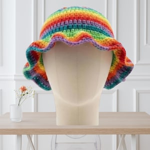 May include: A crochet bucket hat featuring horizontal stripes in a vibrant rainbow color palette including red, orange, yellow, green, blue, and purple. The hat has a rounded crown and a wide, wavy brim with a textured, hand-stitched appearance.
