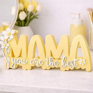 May include: A yellow wooden sign that spells "MAMA" with the phrase "you are the best" in white script. The sign is decorated with white flowers and sits on a white surface. The theme is Mother's Day.