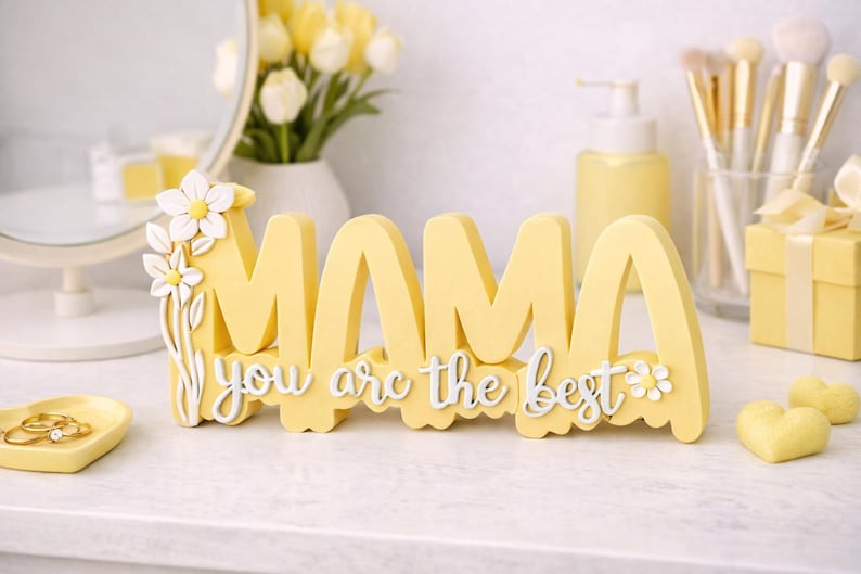 May include: A yellow wooden sign that spells "MAMA" with the phrase "you are the best" in white script. The sign is decorated with white flowers and sits on a white surface. The theme is Mother's Day.