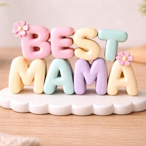 May include: A pastel-colored decorative sign that reads "BEST MAMA". The letters are in pink, yellow, mint green, and lavender, with small flower accents. The sign sits on a white, cloud-shaped base.