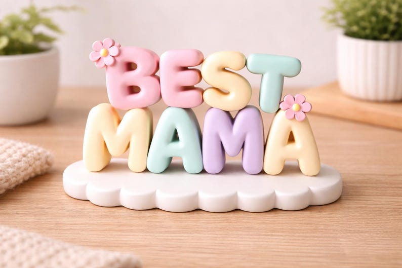 May include: A pastel-colored decorative sign that reads "BEST MAMA". The letters are in pink, yellow, mint green, and lavender, with small flower accents. The sign sits on a white, cloud-shaped base.