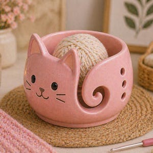 Può includere: Una ciotola per filato in ceramica rosa a forma di gatto, con una palla di filato all'interno. La ciotola ha un design a forma di gatto e un vortice per il filo. Poggia su un tappetino rotondo e intrecciato.