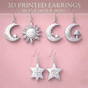 May include: A collection of six silver-toned 3D printed earrings featuring celestial and nature-inspired motifs. The designs include a crescent moon with a star, a sun with a face, a crescent moon with stars, a crescent moon with a mushroom, and two star-shaped pendants, one with a central star and one with an eye symbol.
