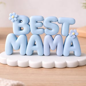 May include: A light blue "BEST MAMA" decorative sign. The letters are in a rounded font and sit on a white cloud-shaped base. Small flower accents are on the letters. The sign is on a wooden surface.