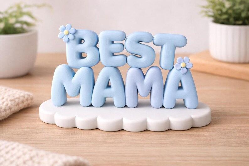 May include: A light blue "BEST MAMA" decorative sign. The letters are in a rounded font and sit on a white cloud-shaped base. Small flower accents are on the letters. The sign is on a wooden surface.