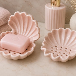 Könnte beinhalten: Zwei hellrosa Seifenschalen aus Keramik in Form von geöffneten Muscheln. Eine Schale enthält ein rechteckiges rosa Seifenstück mit einem kleinen Muschelaufdruck. Beide Schalen haben einen gewellten Rand und einen Boden mit Abflusslöchern.