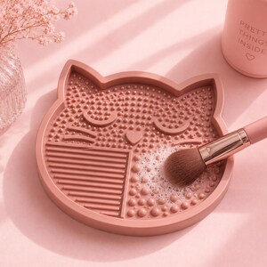 Op de afbeelding: Een roze katvormige make-up borstelreinigingsmat met verschillende getextureerde oppervlakken. Een make-up borstel wordt met zeep en water op de mat gereinigd. Een roze fles met de tekst "PRETTY THINGS INSIDE" staat op de achtergrond.