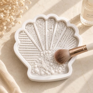 Peut inclure: Un tapis de nettoyage pour pinceaux de maquillage blanc en forme de coquillage avec diverses surfaces de nettoyage texturées. Un pinceau de maquillage avec une virole dorée et un manche blanc repose sur le tapis.