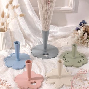May include: A collection of five candle holders in various pastel colors including blue, grey, pink, cream, and sage green. Each holder features a flat, flower-shaped base with a central cylindrical stem and a small heart-shaped cutout detail.
