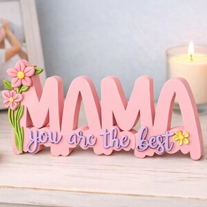 May include: A pink wooden sign that spells out "MAMA" with the phrase "you are the best" in purple script. The sign is decorated with pink flowers and green leaves. A framed photo, flowers, and a candle are in the background.