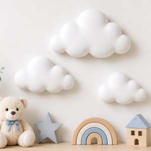 Puede incluir: Tres decoraciones de pared blancas con forma de nube redondeada dispuestas sobre un oso de peluche beige con un lazo azul, una estrella gris, un arcoíris de madera, una casita de madera con techo azul y un jarrón blanco con eucalipto.