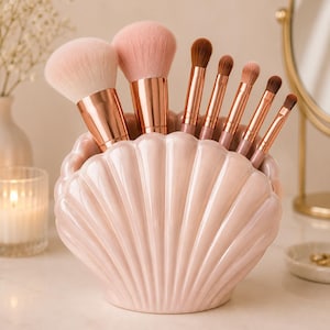 May include: A light pink ceramic shell-shaped holder containing seven makeup brushes with rose gold ferrules and soft bristles. The brushes vary in size, ranging from large fluffy powder brushes to smaller, tapered eyeshadow and blending brushes.