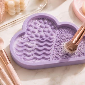 May include: A lavender cloud-shaped makeup brush cleaner with various textured surfaces for cleaning brushes. A makeup brush is being cleaned in the soapy water. Several makeup brushes with pink handles are visible.