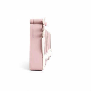 May include: A light pink, decorative wall shelf with a detailed, ornate design. The shelf has a rectangular shape with a series of vertical, white accents. The shelf is designed to be mounted on a wall and is likely used for displaying small items.