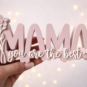 May include: A pink wooden sign that says "MAMA you are the best" in a cursive font. The sign has a floral design on the left side and is held by a hand. The background has soft, blurred lights.