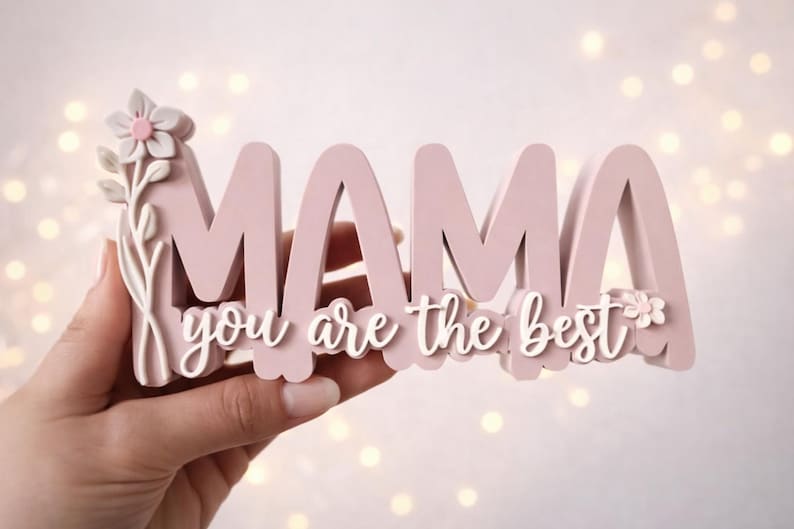May include: A pink wooden sign that says "MAMA you are the best" in a cursive font. The sign has a floral design on the left side and is held by a hand. The background has soft, blurred lights.