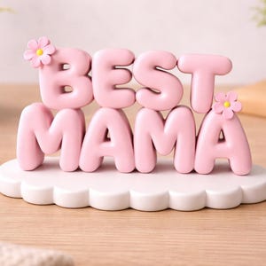 May include: A pink decorative sign that reads "BEST MAMA" in bubble letters. The letters sit on a white, cloud-shaped base and are adorned with small pink flowers. The sign is on a wooden surface.