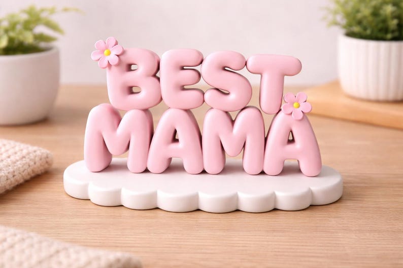 May include: A pink decorative sign that reads "BEST MAMA" in bubble letters. The letters sit on a white, cloud-shaped base and are adorned with small pink flowers. The sign is on a wooden surface.